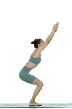 Yeşil elbiseli genç bir kadının beyaz arka planda yoga pozu (utkatasana) verirken çekilmiş bir fotoğraf.