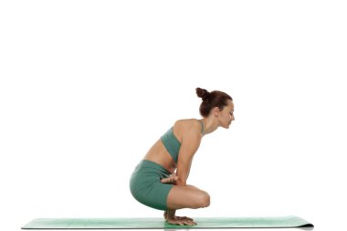 Yeşil elbiseli genç bir kadının beyaz arkaplan üzerine odaklanmış ifadesiyle yoga (tolasana) pozu çalışmasının stüdyo fotoğrafı.