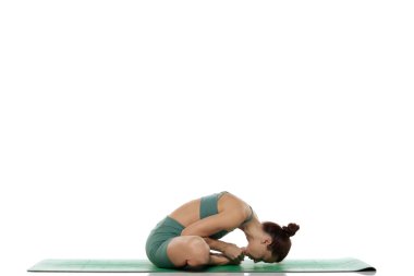 Yeşil kıyafetli genç bir kadının, beyaz arka planda öne eğimli yoga pozu (baddha konasana) çalışırken çekilmiş bir fotoğrafı.