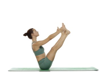 Yeşil elbiseli genç bir kadının, beyaz arka planda yoga pozu (paripurna navasana versiyonu) uygulamasını çekiyoruz.