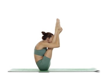 Yeşil elbiseli genç bir kadının yoga yaparken çekilen fotoğrafı. Bacakları kaldırılmış, kolları öne sarılmış, beyaz arka planda bükülmüş.