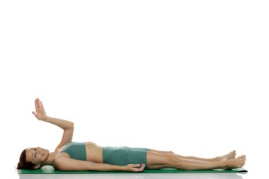 Stüdyoda yeşil elbiseli gülümseyen genç bir kadın var. Savasana pozunda uzanmış, el sallayarak beyaz arka planda.