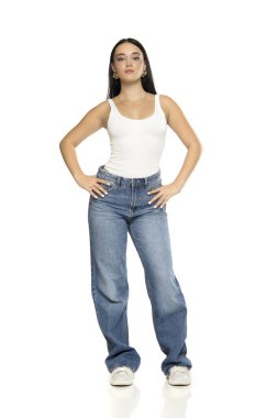 Uzun siyah saçlı, beyaz kolsuz bluzlu, kot pantolonlu ve spor ayakkabılı, elleri kalçasında beyaz arka planda duran genç bir kadının stüdyo fotoğrafı.