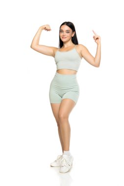 Nane yeşili aktif giyimli, beyaz spor ayakkabılı, kollarını esneten ve beyaz arka planda enerji dolu reklam pozuyla yukarı bakan genç bir kadının stüdyo fotoğrafı.