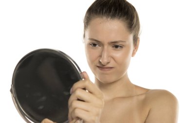 Makyajsız genç bir kadının beyaz arka planda aynada kendine kızgın bakışları.