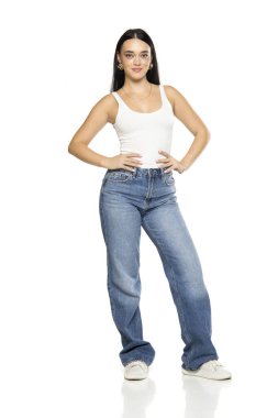 Uzun siyah saçlı, beyaz kolsuz bluzlu, kot pantolonlu ve beyaz arka planda poz veren genç bir kadının stüdyo fotoğrafı.