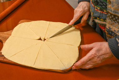 Kadın hamuru ahşap bir tahtanın eşit kısımlarına ayırarak sıcak bir mutfakta pişirmeye hazırlanıyor. Atmosfer bir rahatlık ve geleneği yansıtıyor.