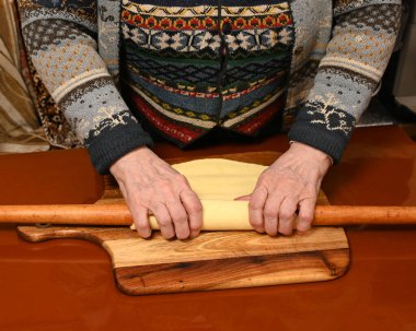 Sıcak dekore edilmiş bir mutfakta tahta bir tahtanın üzerine hamur döken bir kadın. Geleneksel yemek yapma tekniklerini ve ev yapımı yemeklere olan bağlılığı yansıtıyor..