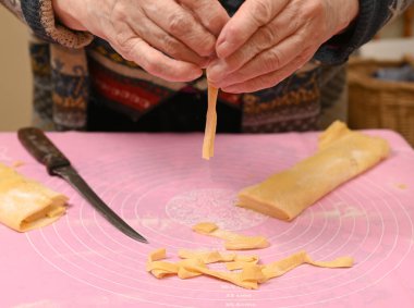 Sıcak bir mutfak ortamında makarnayı ustalıkla şekillendiren ve kesen bir kadın. Tezgah un ve makarna şeritleriyle kaplı. Yemek pişirmeye hazırlanıyorlar.