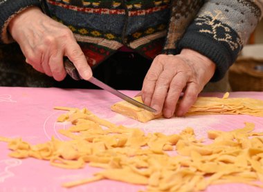 Sıcak bir mutfak ortamında makarnayı ustalıkla şekillendiren ve kesen bir kadın. Tezgah un ve makarna şeritleriyle kaplı. Yemek pişirmeye hazırlanıyorlar.