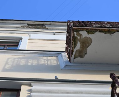 Binanın dış cephesinde mavi gökyüzünün altında soyulmuş boya ve paslı balkon görünüyor.