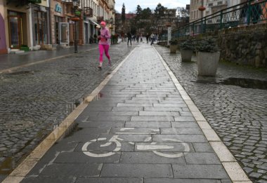 Uzhhorod, Ukrayna - 13 Mart 2025: Parlak idmanlı koşucu kaldırım taşı sokağının yakınındaki bisiklet yolu boyunca koşar. Islak asfalt, insanlar yakınlarda dolaşırken bulutlu gökyüzünü yansıtır.