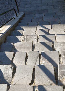 Güneş ışığı, şehrin eski bir bölgesindeki sarp basamaklardan oluşan yıpranmış kaldırım taşları boyunca uzun gölgeler bırakır. Taşların dokusunu ve tarihini vurgular.
