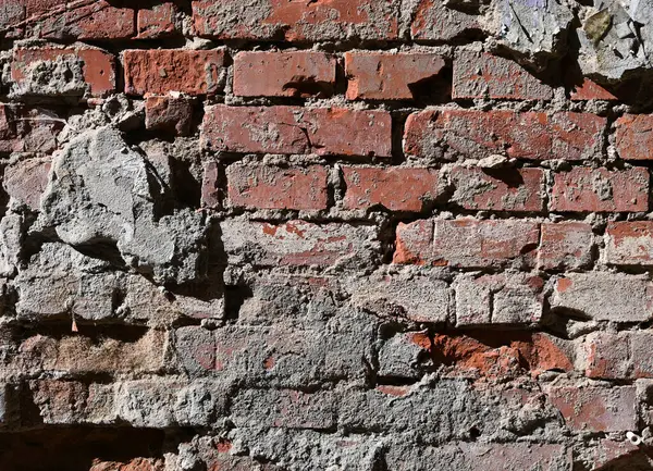 Kırmızı tuğlalar ve gri harç karışımından oluşan yıpranmış tuğla duvarın yakın çekimi. Yüzey çatlakları ve soyulmuş yamaları gösterir, yaşı gösterir
