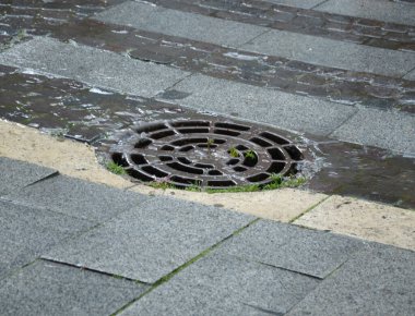 Yağmur durmaksızın yağarken, bir kaldırım taşı caddesinde su, yuvarlak metal bir drenajın etrafında akıyor.