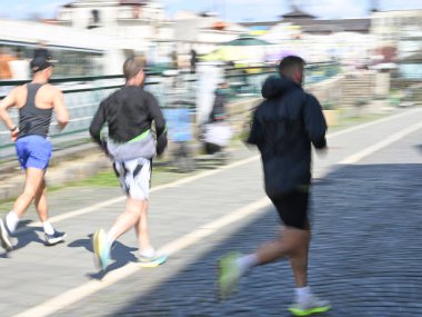 Koşucular güneşli bir günün tadını çıkarıyorlar. Canlı bir şehir ortamında liman boyunca koşuyorlar.