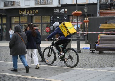 Uzhhorod, Ukrayna - 14 Nisan 2025: Teslimat işçisi, Uzhhorod 'daki çeşitli dükkanların önünde yayaları geçerken kaldırım taşı caddesi boyunca bisiklet sürüyor