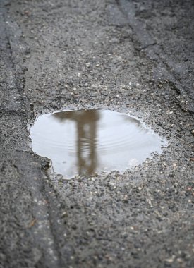 Çatlamış asfalt bir yüzeyde sığ bir su birikintisinde toplanan su, bulutlu bir günde yakınlardaki inşaat malzemelerinin siluetini yansıtıyor.