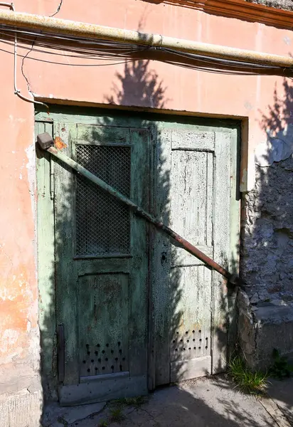 İki yaşlı yeşil çift kapı kısmen kapalı, tahta bir kirişle korunuyor. Etrafındaki duvarlarda aşınma izleri var. Güneş ışığı yakındaki bitkilerin arasından süzülüyor.