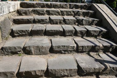 Manzara, güneşli bir parkta yükselen pürüzlü taşlardan yapılmış bir dizi düz olmayan taş basamakları gösteriyor.