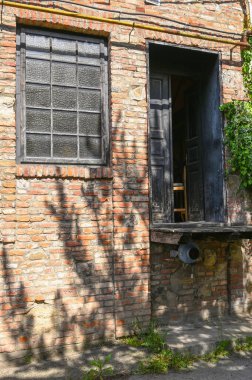 Kırsal tuğla yapı, açık ahşap kapılar ve bir balkona sahiptir. Ağaçların gölgeleri, sakin bir ara sokakta yüzeyi hafifçe aydınlatır. 