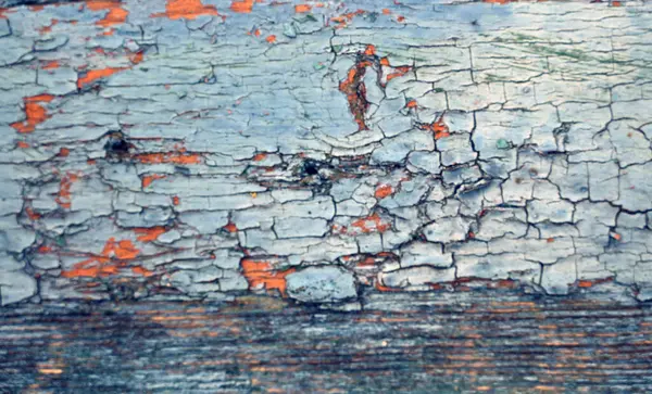 Yakından bakınca, çatlamış boyayla yıpranmış ahşap bir yüzey görünüyor. Mavi ve turuncu renkler sergiliyor. Doku ve renkler, yaşlanan bir yapının kırsal cazibesini vurgular.