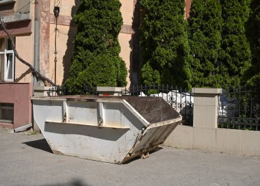 Büyük, boş bir çöp konteynırı düzgünce budanmış yeşilliklerle çevrili bir binanın yanındaki kaldırıma yerleştirilmiş.