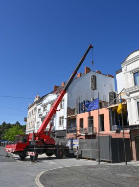 Uzhhorod, Ukrayna - 12 Mayıs 2025: Kırmızı vinç bir şehir caddesinde, inşaat malzemelerini bir şantiyeye taşır. Açık mavi gökyüzü kentsel gelişimin canlı görüntüsünü artırıyor