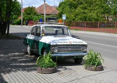 Uzhhorod, Ukrayna - 12 Mayıs 2025: Yeşil ve beyaza boyanmış klasik polis arabası, sakin bir caddenin kenarına park edilmiştir. Bitkilerle çevrili saksılar açık mavi gökyüzünün altında sahneye cazibe katar.