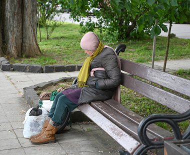 Uzhhorod, Ukrayna - 12 Mayıs 2025: Kadın ahşap bir park bankında oturuyor, başörtüsü ve eşarbı ile kalın giysiler içinde. Eşyaları çantaların içinde dinleniyor gibi görünüyor.