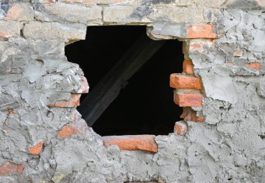 Yıpranmış tuğla duvarda belirgin bir delik, arkasındaki ahşap kirişleri ortaya çıkarıyor. Yapı, doğal ortamdaki kırık tuğlalar ve pürüzlü alçıyla yaş belirtileri gösteriyor.