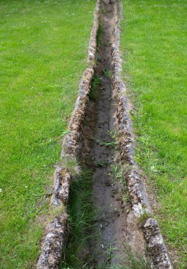 Uzun, dar kanalizasyon kanalı yemyeşil bir alanda uzanıyor. Yaşlanmış taş kenarlar kısmen çimen ve toprakla kaplıdır. Bu da yıllar süren doğal aşınmayı gösterir.