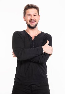 Sakallı genç adam, kolları bağlı, siyah uzun bir gömlek giyiyor ve dostça bir gülümseme sergiliyor.