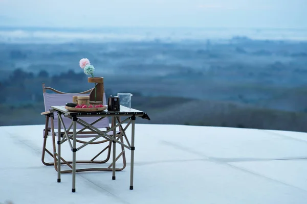Empty outdoor chair and set of coffee cups on the folding table with blurred landscape with fog in background