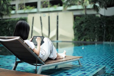Selective focus on hand of woman sits on the bench and writes  on the screen by the bluetooth pen with blurred swimming pool in background 