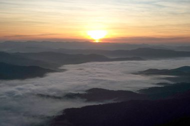 Panorama view layers of mountain with the fluffy white fog between the mountains and orange sunrise sky in background clipart