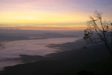Dağlar arasında kabarık beyaz sis ve arka planda turuncu gün doğumu gökyüzü ile Panorama manzaralı dağ katmanları
