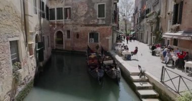VENICE, İtalya - 10 Nisan 2023: iki gondollu dar bir deniz kanalı ve insanlar Calle Larga adlı küçük bir ara sokakta, romantik ve huzurlu bir manzaranın restoranlarında oturuyorlar..