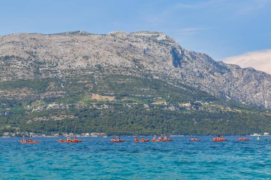 KORCULA, CROATIA - 24 Haziran 2019: Turistli bir grup kano Peljesac yarımadası ve St. Ilija Dağı ile birlikte arka planda süzülüyor.