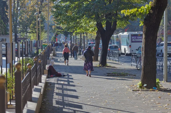 stock image TIRANA, ALBANIA - October 20, 2019: Pedestrians walk on a wide city sidewalk while a beggar woman sits by the wall seeking alms.