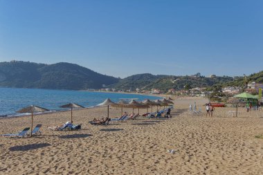 CORFU, GREECE - 21 Ekim 2022: Ziyaretçiler, arka planda yemyeşil tepeler bulunan Korfu 'daki kumlu bir plajda saman şemsiyeler altında güneşlenirler..