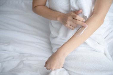 Asyalı kadın yatakta oturuyor ve vücuduna krem sürüyor..
