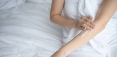 Asyalı kadın yatakta oturuyor ve vücuduna krem sürüyor..