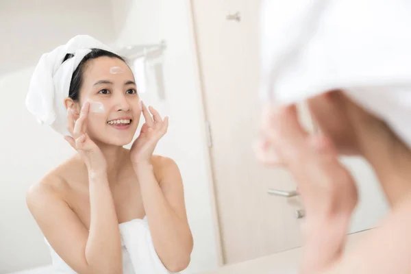 快乐的年轻亚洲女人穿着毛巾 一边擦着脸一边涂面部乳液 — 图库照片