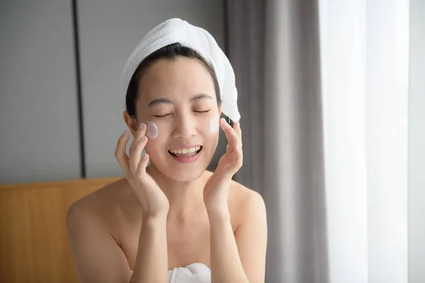 Happy young Asian woman applying face lotions while wearing a towel and touching her face.