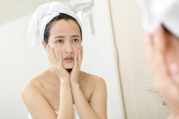 Problem skin. Concerned young asian women popping pimple on cheek while standing near mirror in bathroom. Unhappy girl inspecting face, suffering acne, focus on reflection