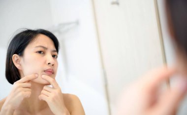 Sorunlu cilt. Endişeli genç Asyalı kadınlar banyoda aynanın yanında dikilirken yanaklarına sivilce çıkarıyorlar. Mutsuz kız yüzünü yokluyor, sivilce çekiyor, yansımaya odaklanıyor.