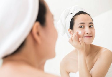 Mutlu genç Asyalı kadın havluyu takıp yüzüne dokunurken yüz losyonu sürüyor..