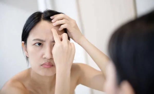 Problem skin. Concerned young asian women popping pimple on cheek while standing near mirror in bathroom. Unhappy girl inspecting face, suffering acne, focus on reflection