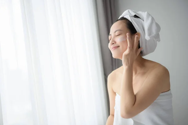 Happy young Asian woman applying face lotions while wearing a towel and touching her face.